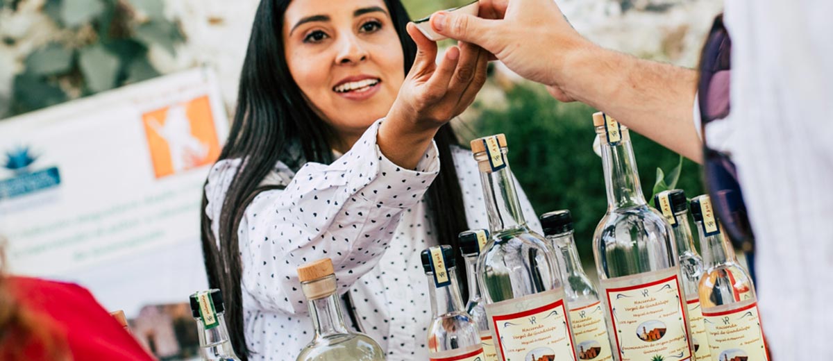 Mujer bartender repartiendo shots en una cata, rodeada de botellas de mezcal